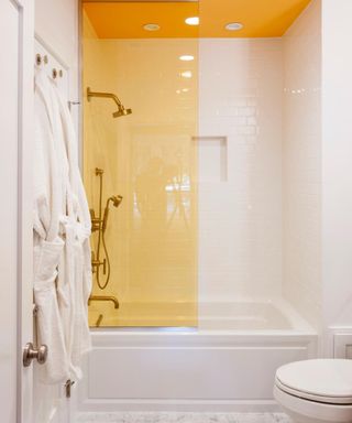 A bathroom with a yellow ceiling and shower screen, a shower bath, white robes on the wall, and a toilet