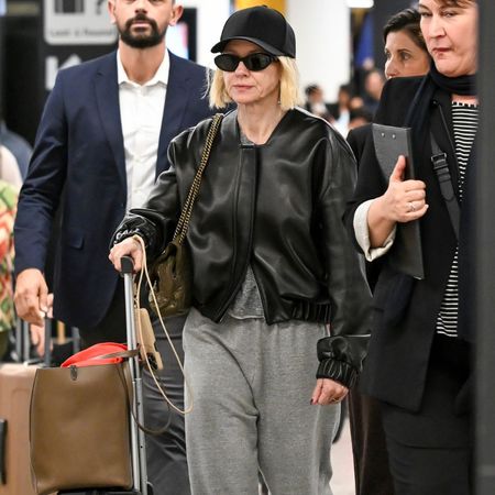 Naomi Watts walks through the airport wearing grey track pants with a black leather bomber jacket, red trainers, a black cap and black sunglasses.