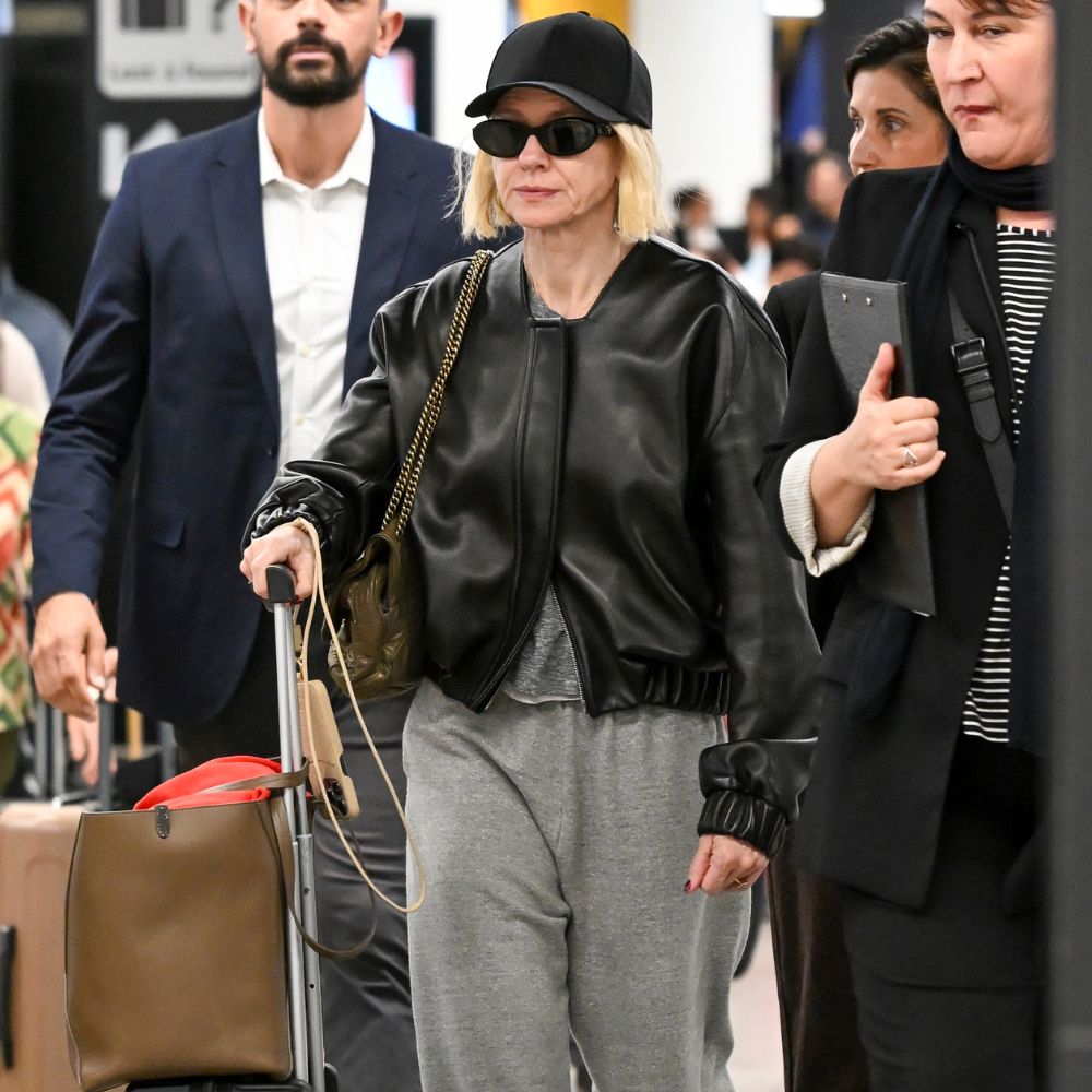 Naomi Watts walks through the airport wearing grey track pants with a black leather bomber jacket, red trainers, a black cap and black sunglasses.