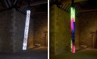 The art installation is in a shape of a long beam with electronic neon writing on it. It's vertical and goes from the ceiling to the floor. There are words and numbers in neon lights.
