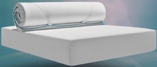 The Simba Cool Foam Mattress Topper rolled up and partially unrolled on top of a white mattress against a soft teal and pink gradient background.