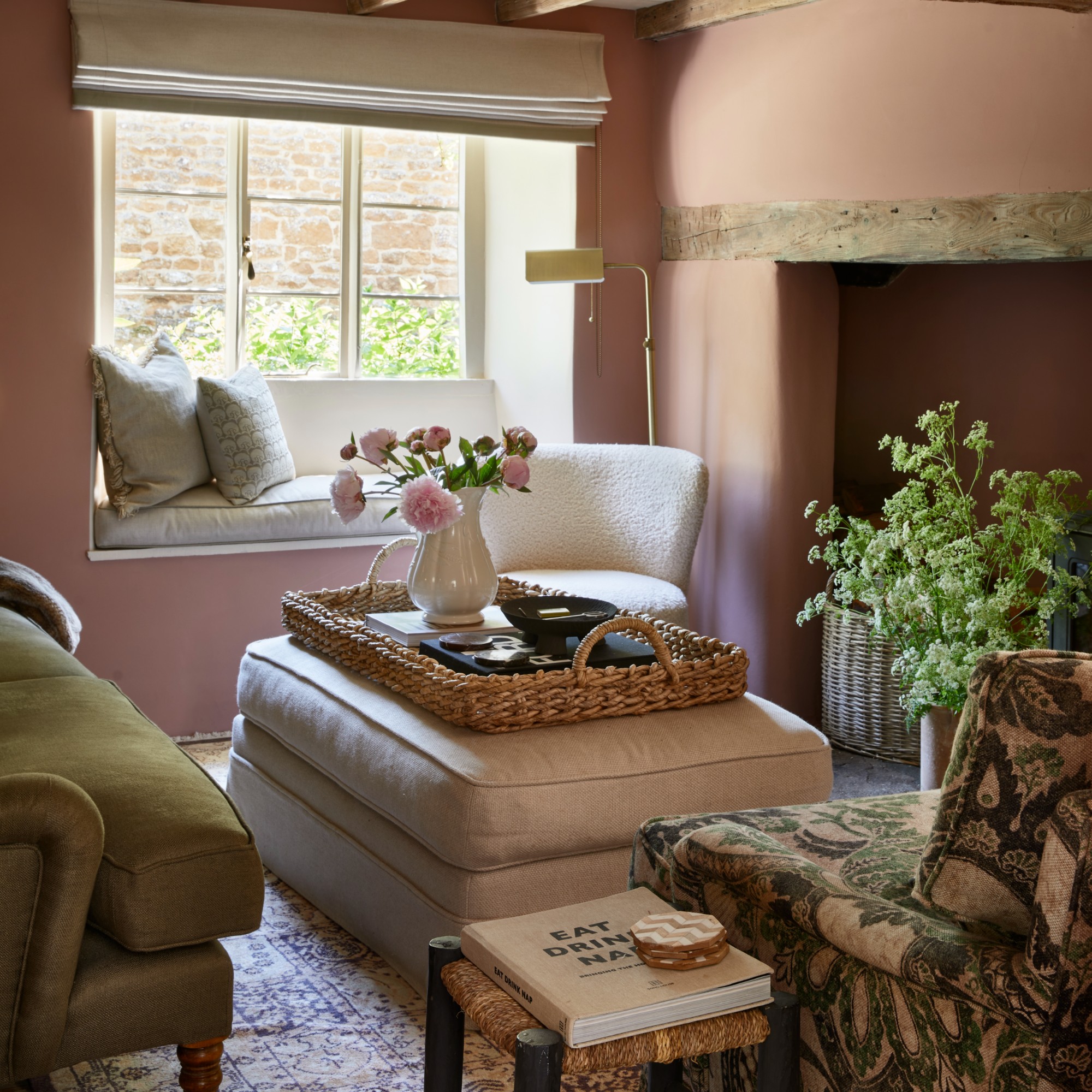 A rustic cottage living room painted pink with a cream storage ottoman in the middle of the seating area used as a coffee table with a rattan tray on top