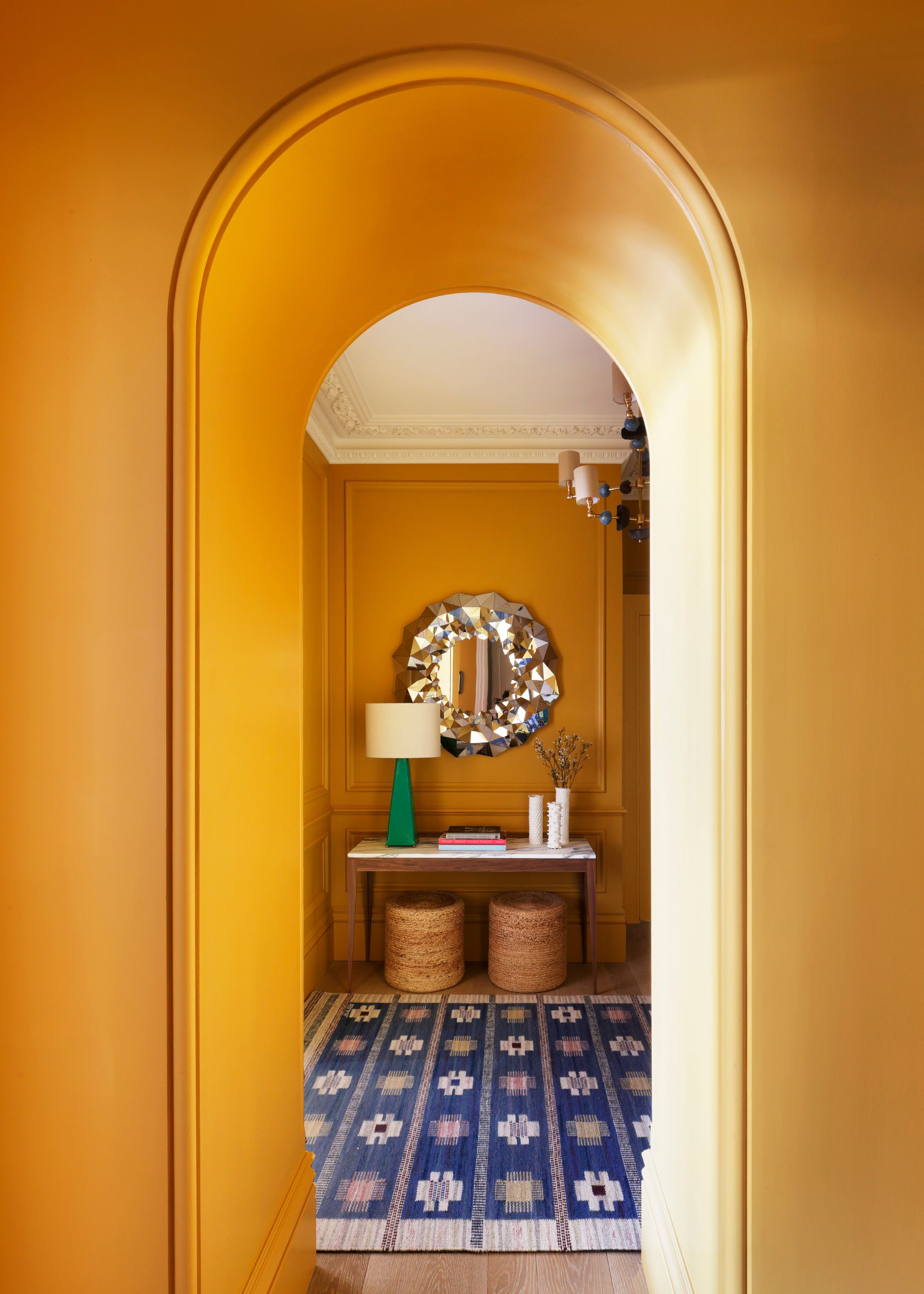 A hallway with an arched doorway painted with a warm shade of ochre. A blue-patterned rug on the ground and a console table with decorative objects and a lamp.