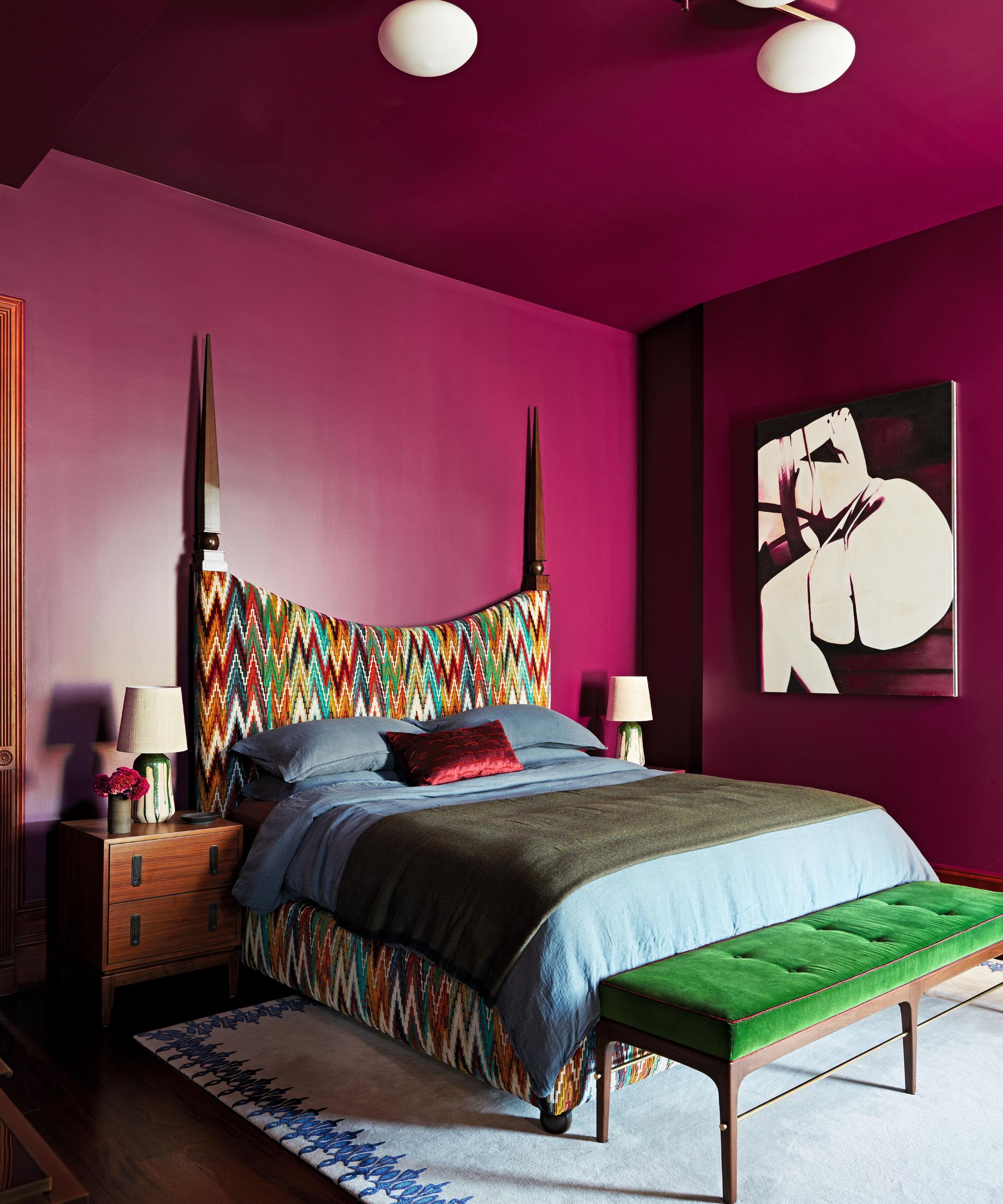 a bright pink purple painted bedroom with statement art and a printed wooden headboard, green end of bed bench and a neutral rug with blue trim detail
