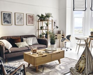 An open plan living room by Homesense with three framed wall art prints, grey velvet upholstered sofa, wooden rectangular sofa, modular shelf which zones office area and dog in teepee