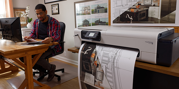 A Canon XL printer sits in the foreground, printing a large architectural document, as a man works at a desk in the background.