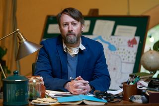 David Mitchell sits at his desk inside the tent, holding a pen in his clasped hands. There is a map displayed on a board behind him, and on the messy desk in front of him are multiple items including binoculars, a wooden duck, an upturned book, a pot of pencils and a plate of biscuits