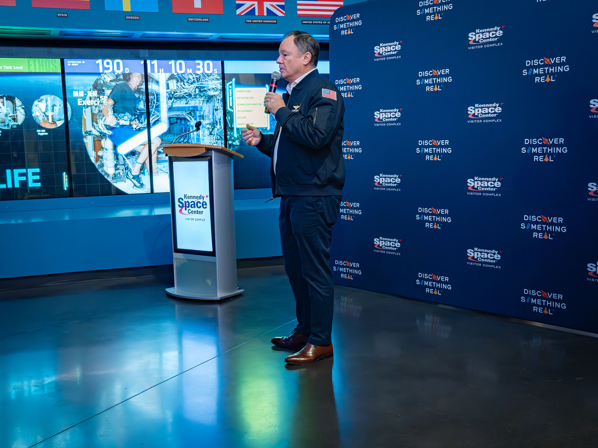 a man in a blue suit speaks with a microphone at an air and space museum