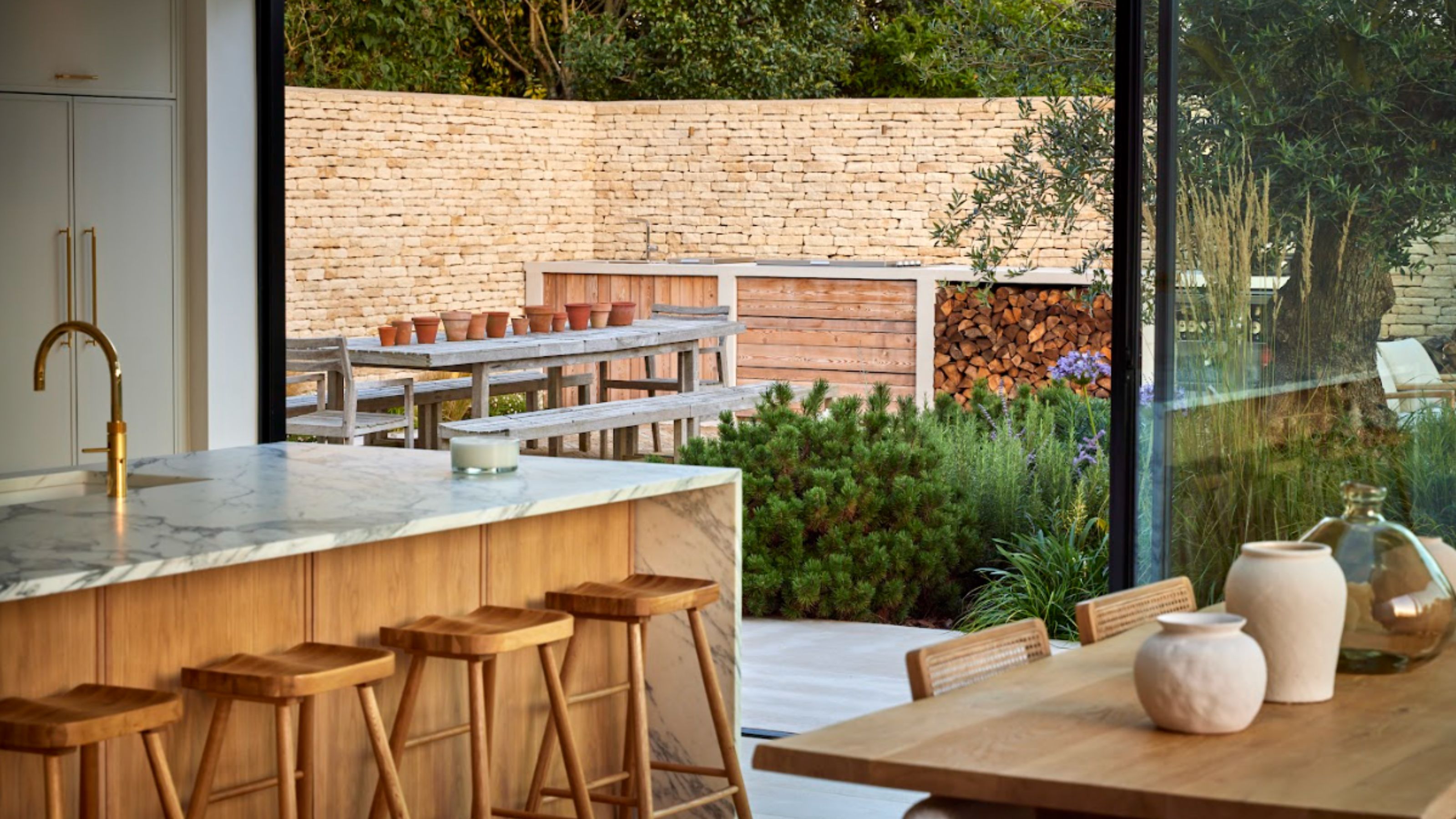 A kitchen with a marble-topped kitchen island with wooden panels and bar stools next to a wooden kitchen table and in front of open sliding glass doors leading to an outdoor kitchen and seating