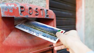 Somebody posting a postcard, with a picture of a tropical beach scene, into a red letterbox