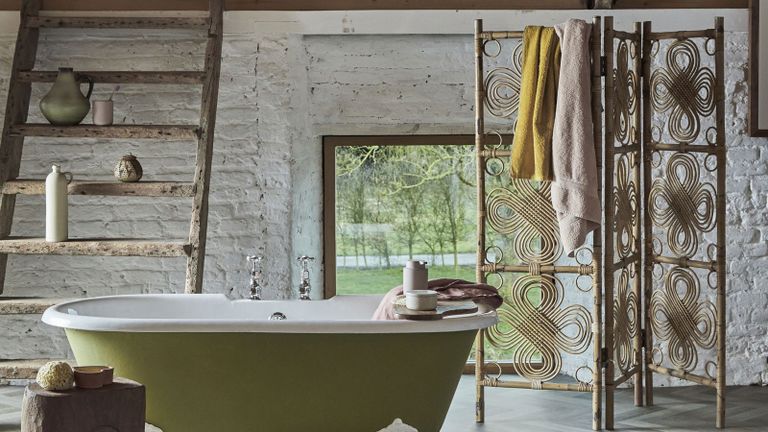 A modern rustic bathroom with green bathtub, herringbone flooring and wooden room divider used as towel storage