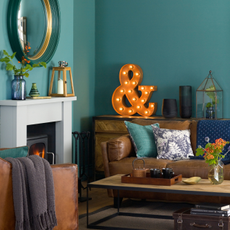 Teal green living room, black woodburner with white mantelpiece, brown leather sofa and amrchair, wood coffee table, large letter light, circular mirror.
