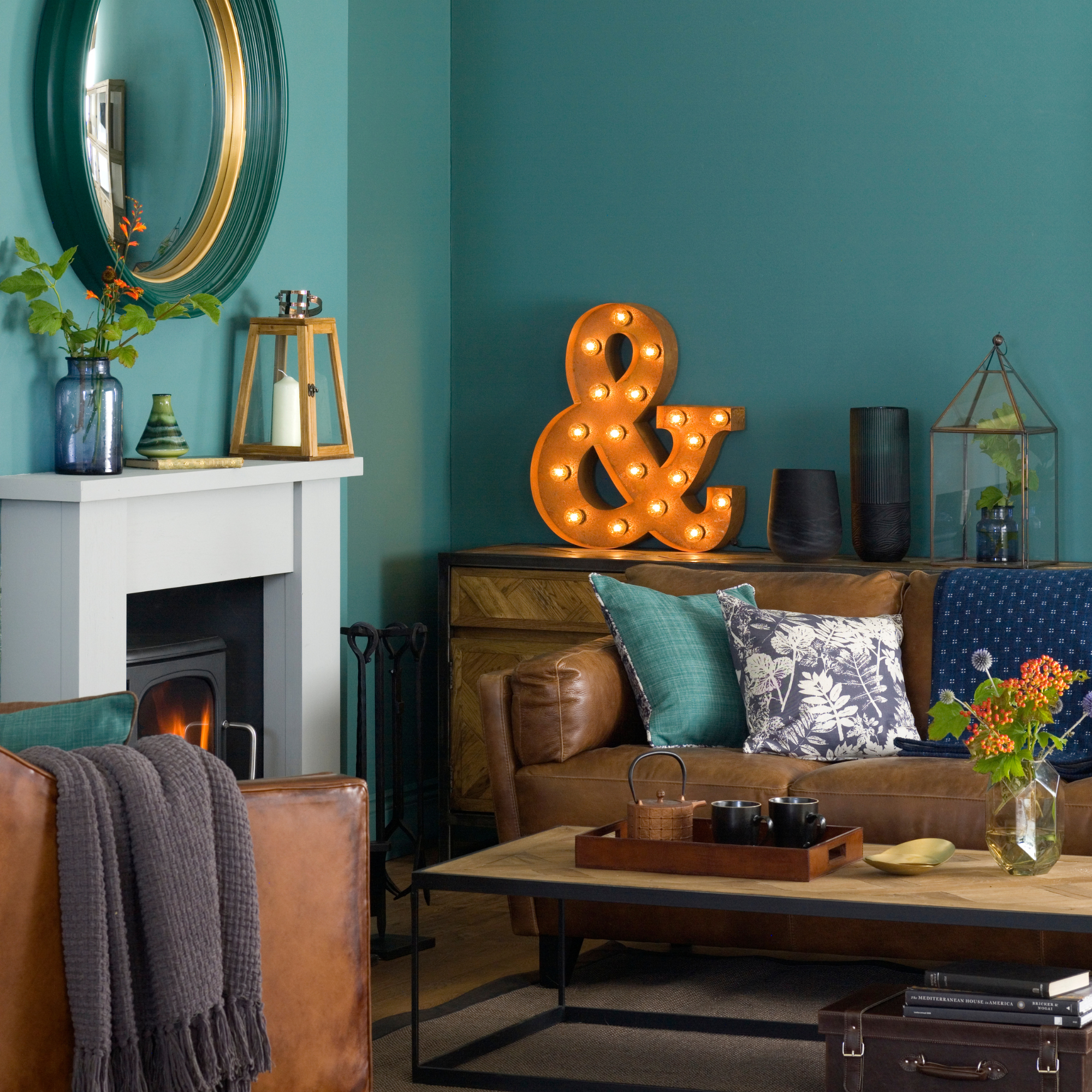 Teal green living room, black woodburner with white mantelpiece, brown leather sofa and amrchair, wood coffee table, large letter light, circular mirror.