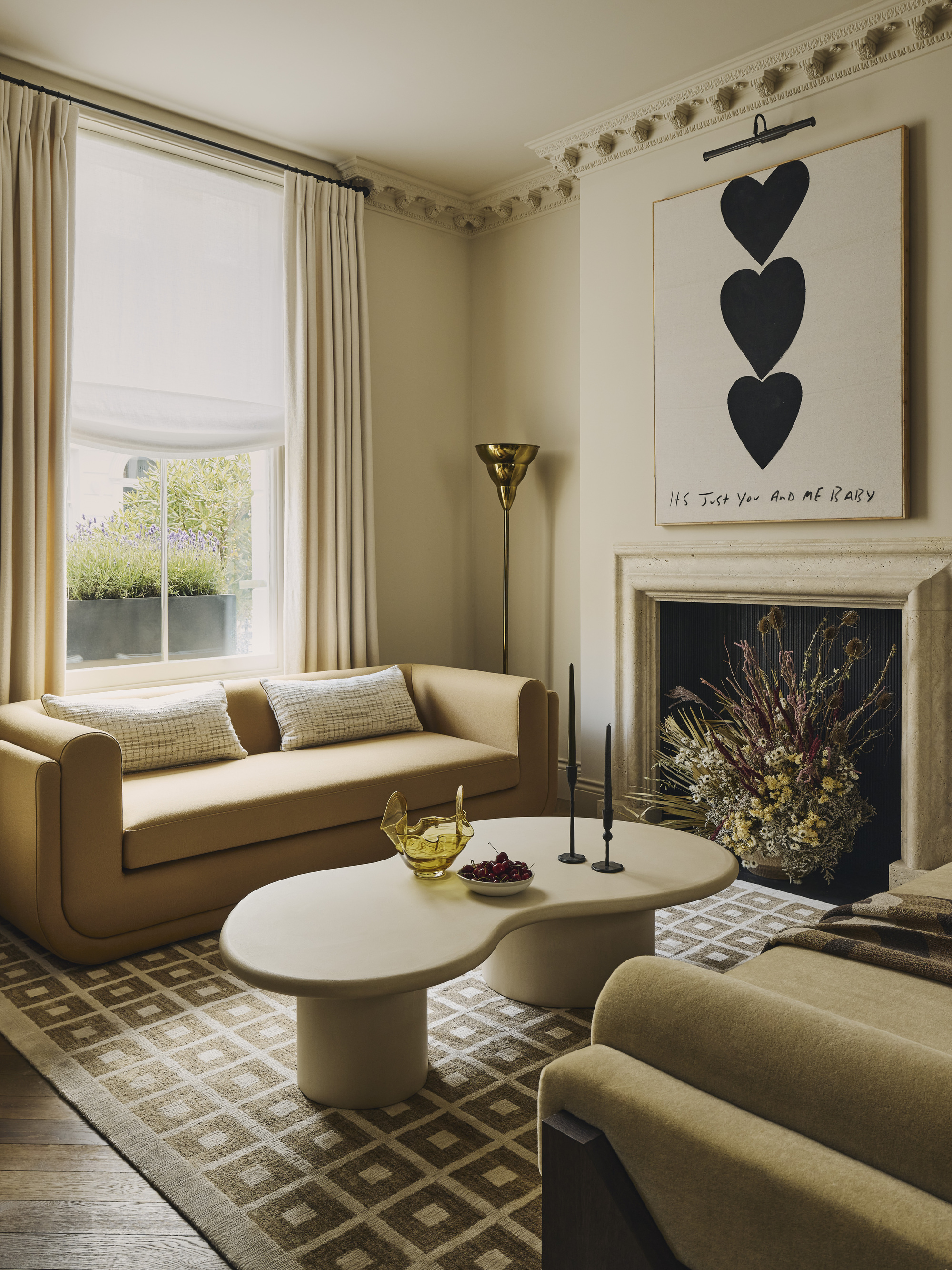 living room with ceiling cornice, layered curtains with sheer roman blinds and cream curtains, beige sofas facing each other, curvy white coffee table in front of fireplace styled with dried flowers with artwork hanging above, and checkered rug