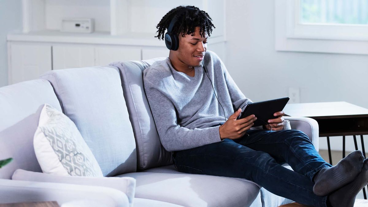 A young Black man watches content on a tablet while sitting on a couch with his feet up.