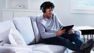 A young Black man watches content on a tablet while sitting on a couch with his feet up.
