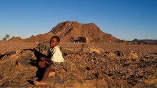 A Nama girl in Northern Cape province, South Africa. She wears a white shirt and a black skirt with white spots. She sits on the rocky ground in front of a small mountain in the background.