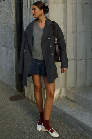 A woman wearing a gray peacoat over a gray v-neck sweater styled with a denim miniskirt, red socks, and white flats.