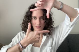 A curly-haired woman in a white shirt frames her own face with her fingers in a mock viewfinder gesture.
