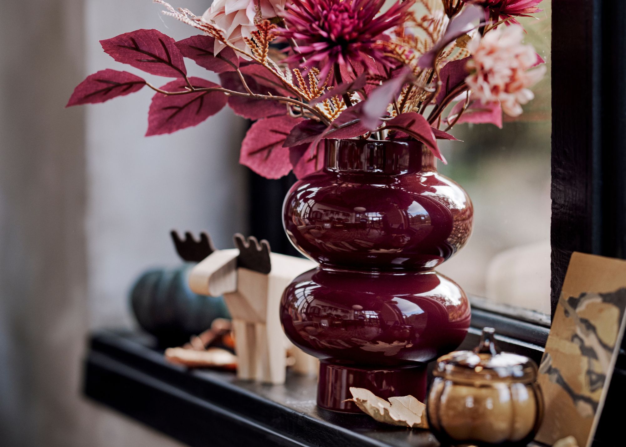 A black windowsill with a glossy brown-red vase with rounded, contemporary shapes styled with autumnal flowers and among other autumnal decor