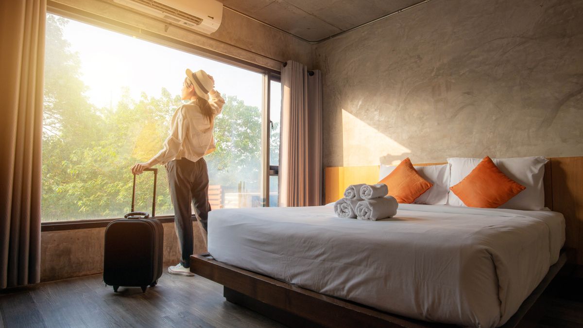 A women wearing a hat and pulling a suitcase looking out of the window in a hotel room