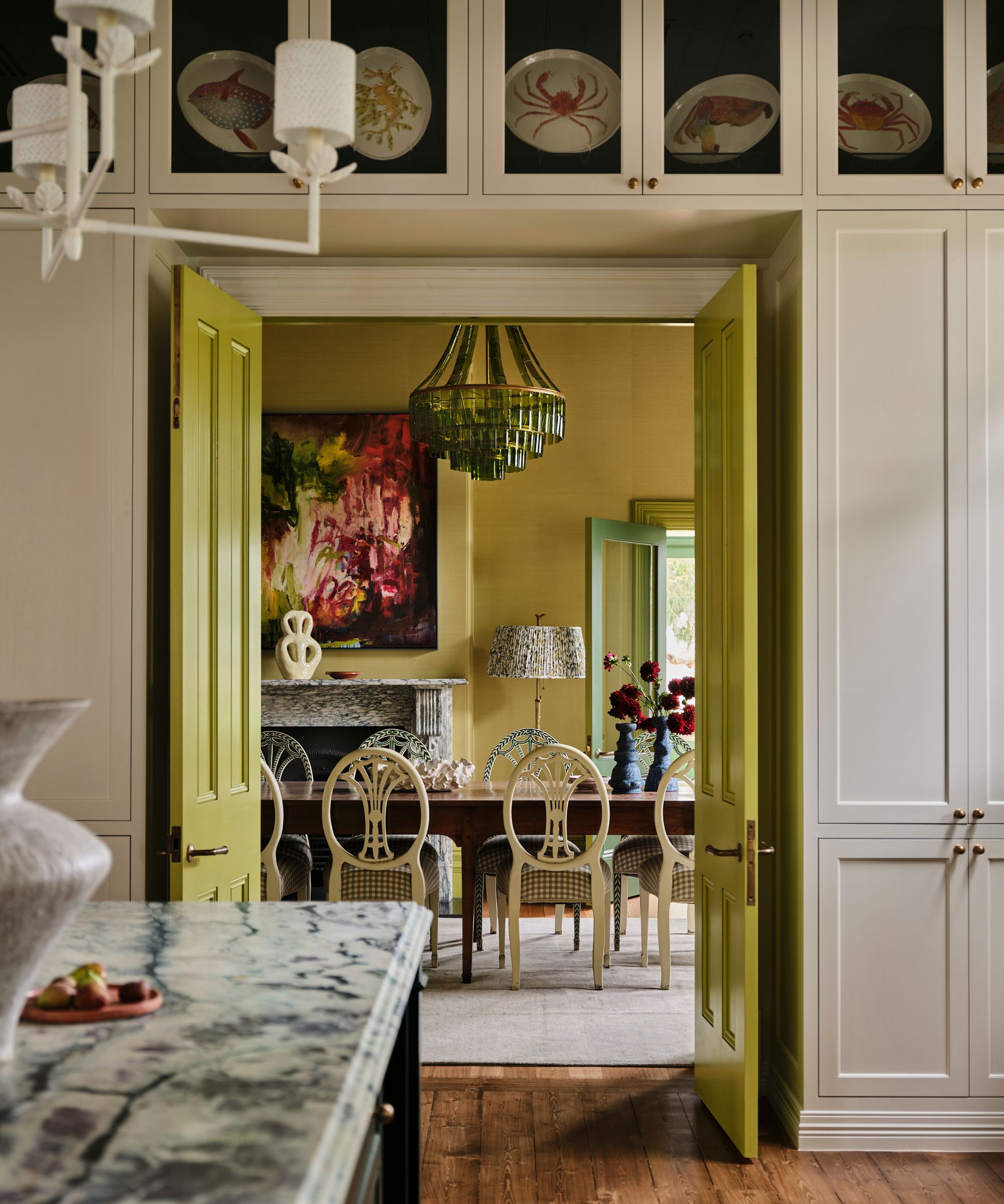 Grand kitchen with marble island, white built-in cabinetry with plates, looking through into the dining room with chartreuse painted doors, green walls, green glass chandelier and long dining table with white chairs