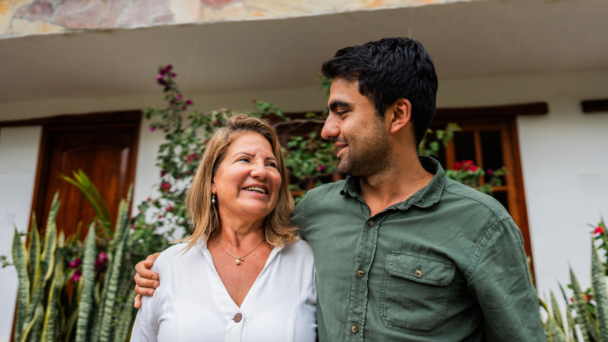 A mother and her adult son smile with each other outside a house. 