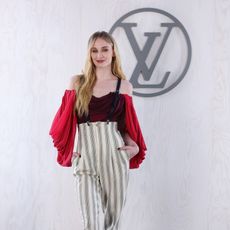 Sophie Turner at Louis Vuitton's PFW show wearing striped suspender pant with a red draped blousents with a 