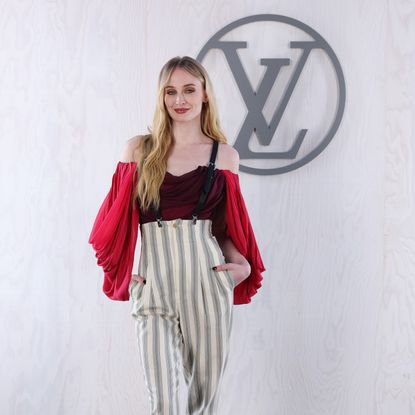 Sophie Turner at Louis Vuitton's PFW show wearing striped suspender pant with a red draped blousents with a 