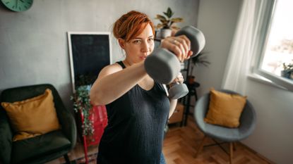 woman wearing a black vest with a living room background holding dumbbells and punching towards the camera 