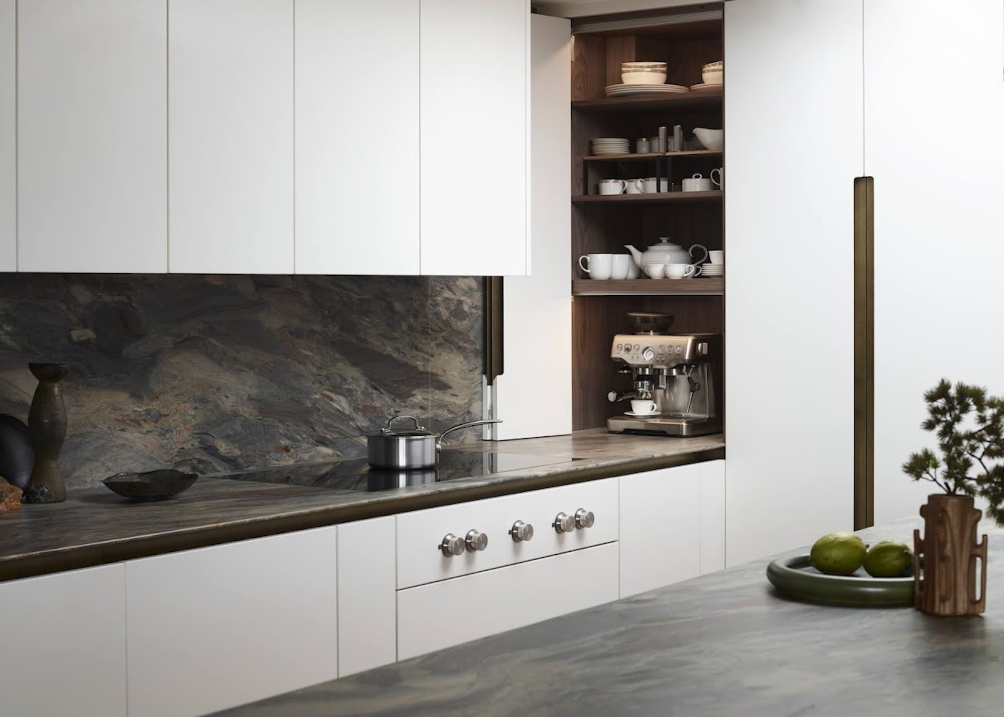 A handle-free white kitchen with striking dark stone worktops and splashback showing a countertop corner cabinet with bi fold doors