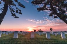 Fort Rosecrans National Cemetery, Point Loma, San Diego, California, USA. Monument headstones along the coast during a pink, orange, and blue sunset with clouds and the ocean in the background and trees, grass, and tombstones in the foreground.