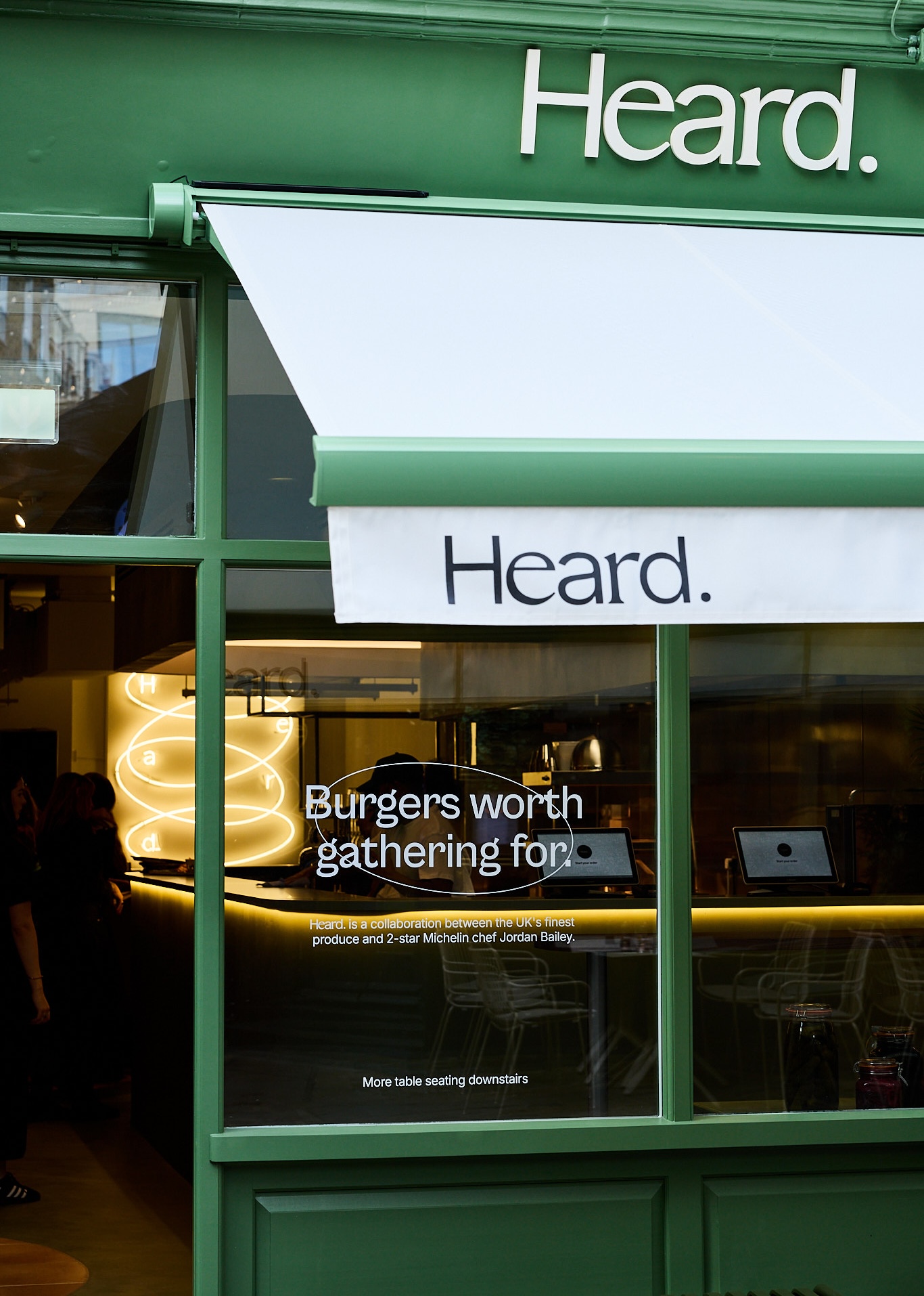 An emerald green restaurant facade features the sign &quot;Heard.&quot; in white as well as in black on white awnings, and a stick-on message on glass that reads &quot;Burgers worth gathering for.&quot; in a circle.