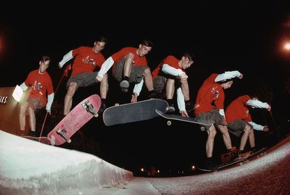 Night time image of a young male skateboarder, red and white long sleeve top, grey shorts, black socks and trainers, photograph is snapped multiple time frames to show the skateboarders movements across the jump, dark sky, street lights