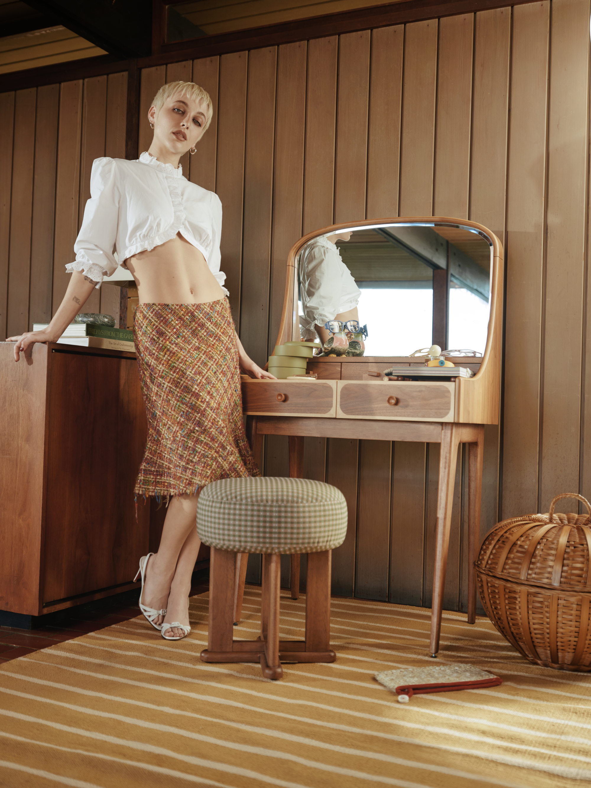 Image of a woman in a cropped white blouse and plaid midi skirt standing next to a mid-century-inspired vanity set in a wood-paneled room.
