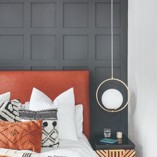 A bedroom with a panelled dark grey wall behind a rust-coloured headboard with a pendant light next to the bed