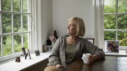 An older woman sits at her table with a coffee cup and looks thoughtfully out the window.