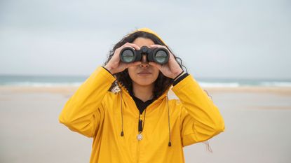 A woman in a yellow raincoat looks through a pair of binoculars.