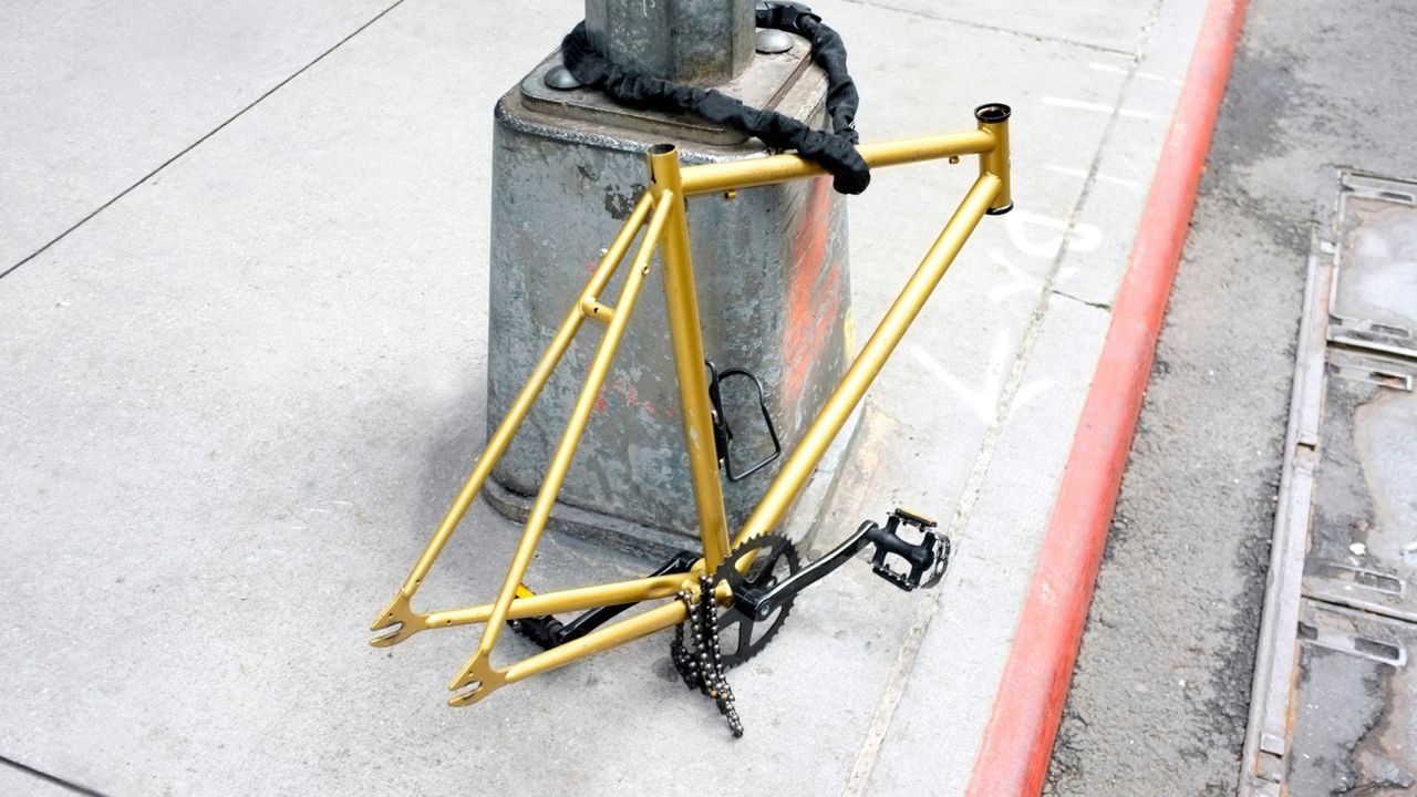 A gold bike frame with missing finishing kit and wheels locked against a post on a pavement