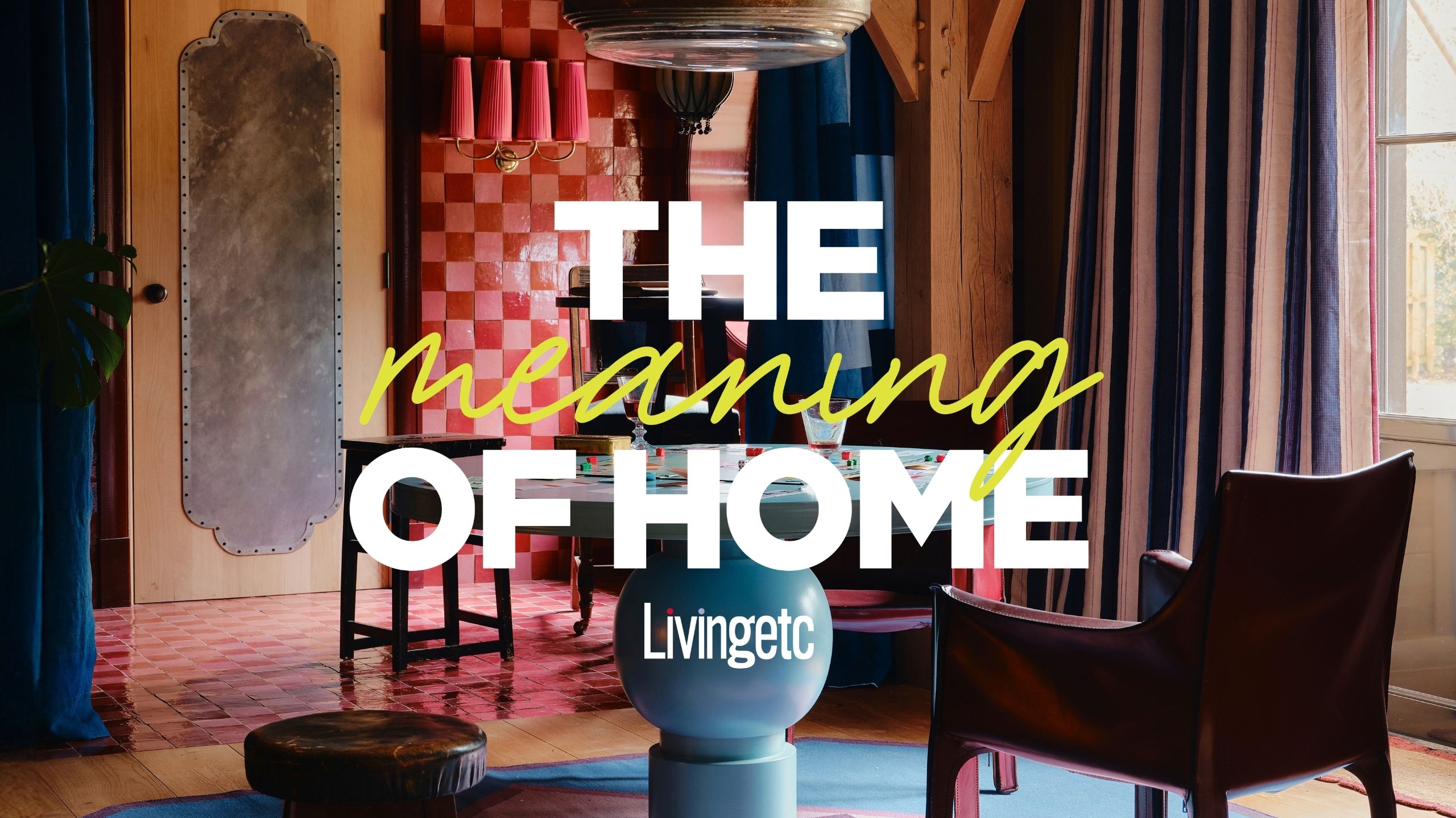 A colorful living room with a lacquered pale blue round table, mismatched leather chairs, a checkerboard wall in tones of pink, and pale blue-washed wooden beams, overlaid with "meaning of home" logo