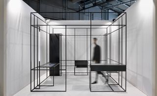 A white space containing an office made out of a square black metal wire frame with a man walking inside of it.