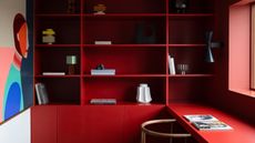 a home office with bright red cabinetry, decor displayed on shelves, timber chair pushed into desk with two magazines, and an artwork on the opposite wall