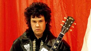 Gary Moore holding a guitar against a red background in 1988