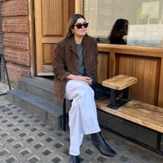 Influencer @theannaedit sits on a bench outside wearing a brown suede blazers with a grey jumper and black square-toe boots.