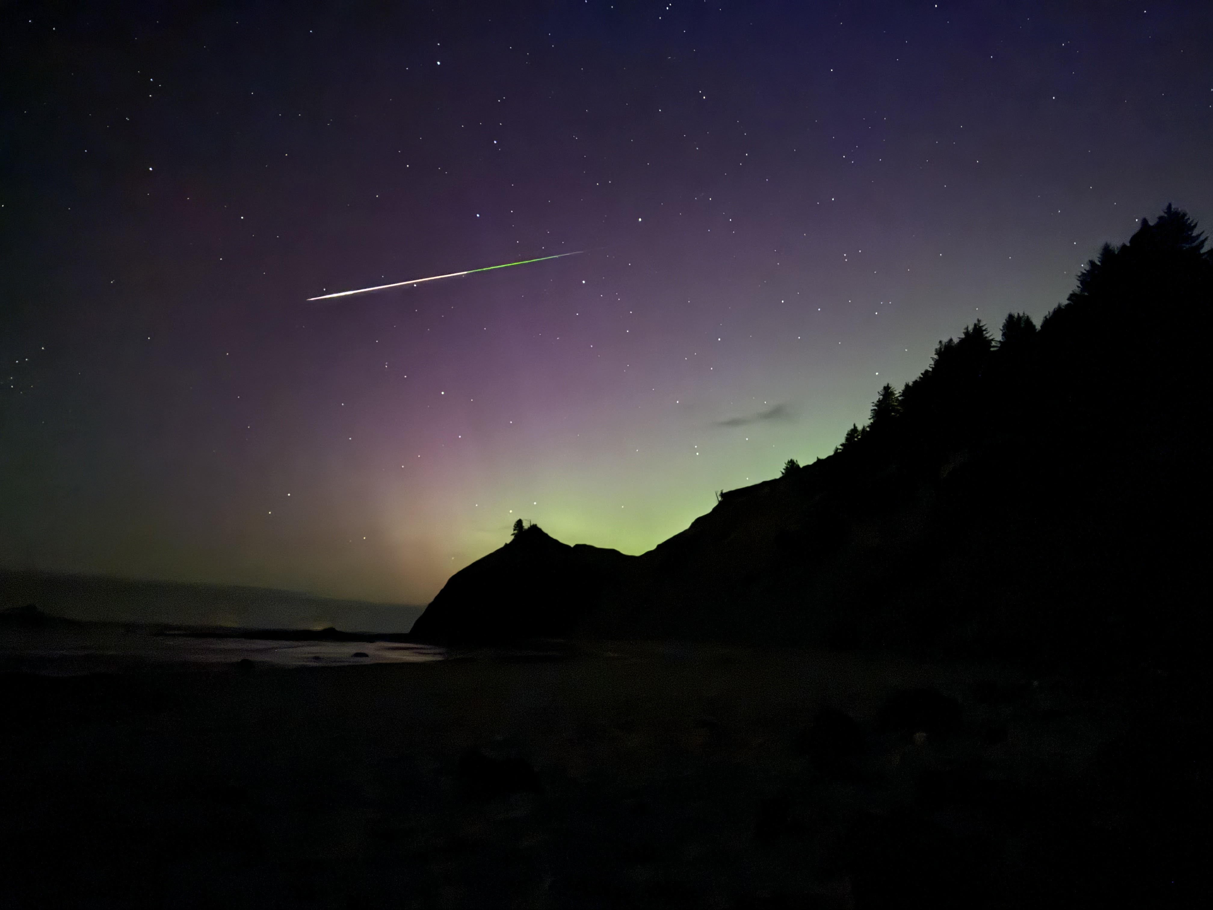 Ein langer, bunter Meteorstreifen verläuft über den Himmel mit einem deutlichen grünen Schimmer gegen Ende des Schweifs, und gleichzeitig leuchtet der Himmel während der Polarlichtdarstellung grün und violett.