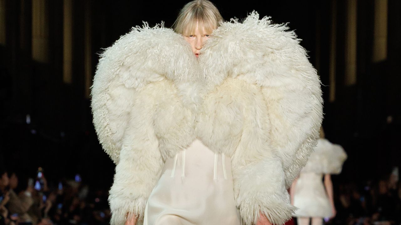 A model wearing a shearling and silk cream-colored coat and dress walking at the F/W 25 Alexander McQueen show.