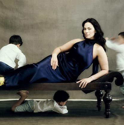 Ashley Graham wears a blue satin gown lying on a couch as her three sons run around her 