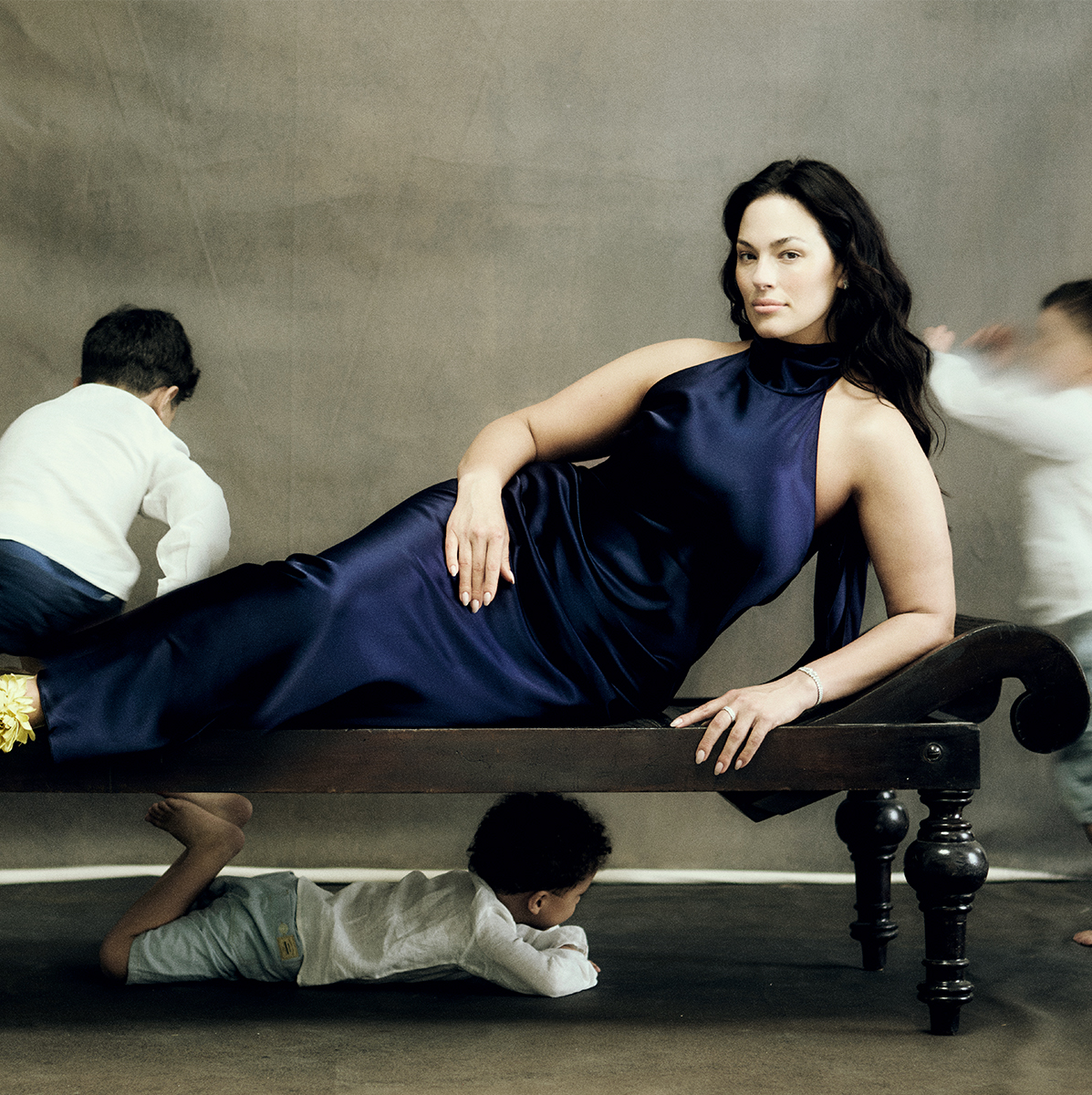 Ashley Graham wears a blue satin gown lying on a couch as her three sons run around her 