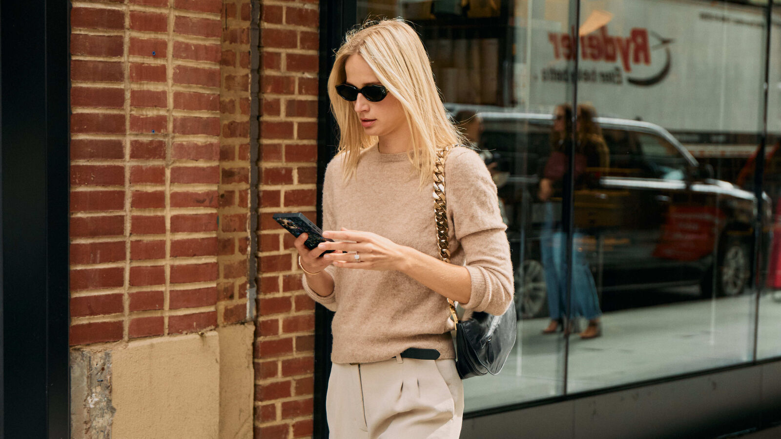 Woman in khaki trousers, beige short-sleeve sweater, sunglasses, and a chain-strap bag 
