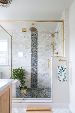 A shower with stone mosaic tiling, a bamboo bath mat, and gold hardware accents